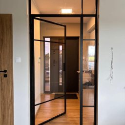 MANAGEMENT GROUP SPÓŁKA Z OGRANICZONĄ ODPOWIEDZIALNOŚCIĄ - Nowoczesna, przeszklona ścianka działowa z czarną ramą, z otwartymi drzwiami, oddziela przestrzeń w domu. Widoczna podłoga z paneli i fragment ściany z kablem.