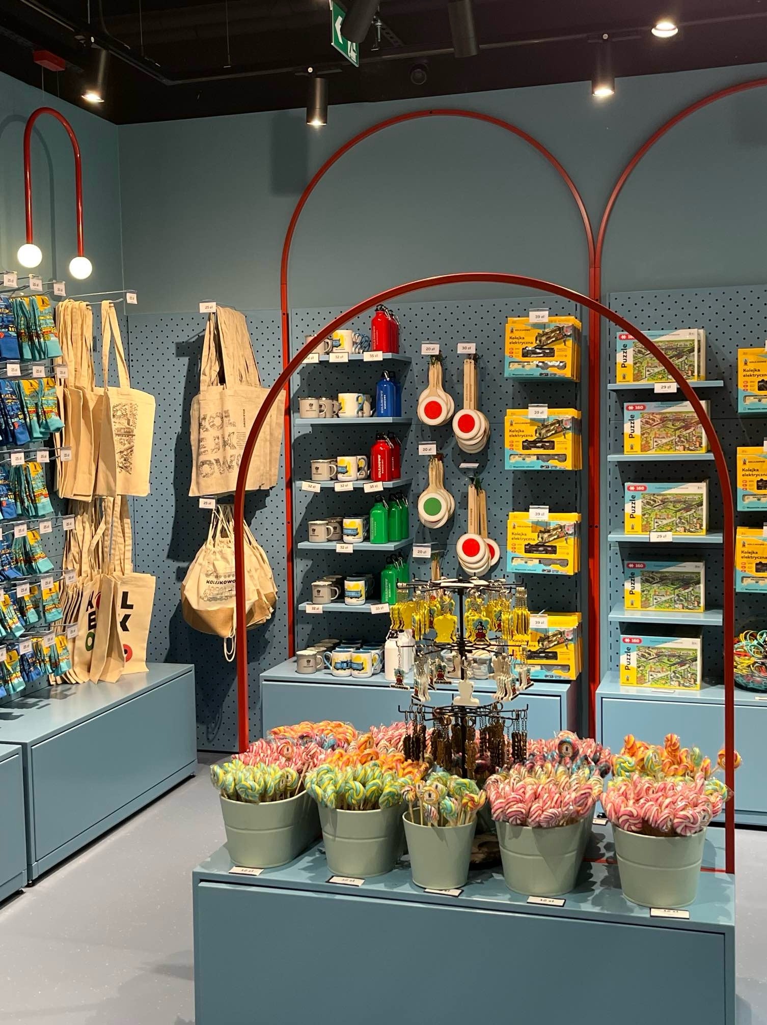 Aranżacja wnętrza sklepu z pamiątkami: niebieskie ściany z czerwonymi łukami, półki z torbami, kubkami, puzzlami i lizakami w wiaderkach.