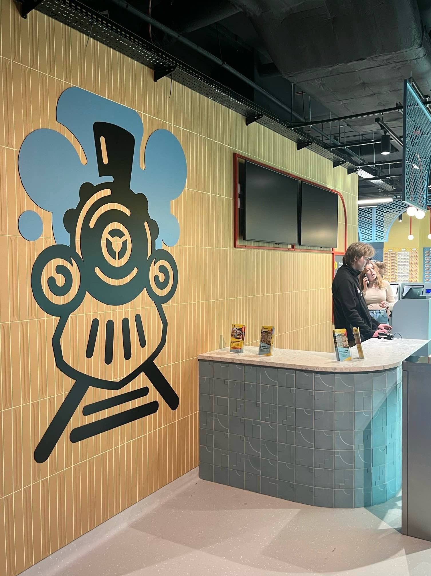 Recepcja z dekoracyjną grafiką lokomotywy na ścianie z beżowych płytek. Za ladą widoczni klienci.