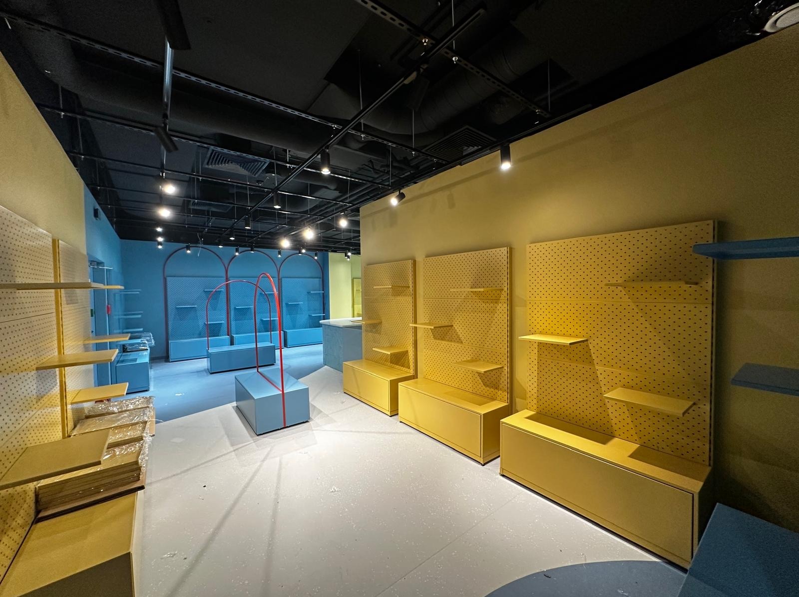 Wnętrze sklepu w trakcie aranżacji: żółte regały i szafki na tle żółtej ściany, niebieskie elementy ekspozycyjne i ściany w tle, czarny sufit z oświetleniem.