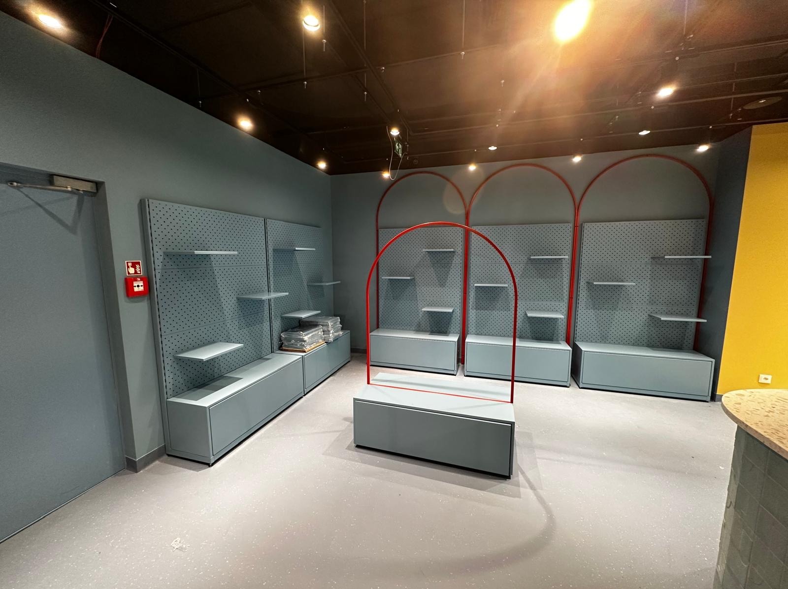 Jasne, minimalistyczne wnętrze sklepu z modułowymi regałami na tle szarej ściany z otworami i czerwonymi, łukowatymi elementami dekoracyjnymi. Nowoczesny design.