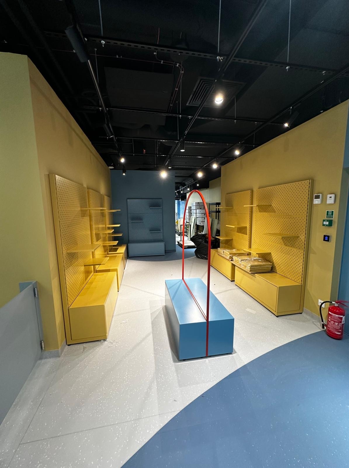 Jasne wnętrze sklepu odzieżowego w trakcie aranżacji: żółte regały z perforowanymi ściankami, niebieska lada ekspozycyjna z czerwoną ramą, szara posadzka.