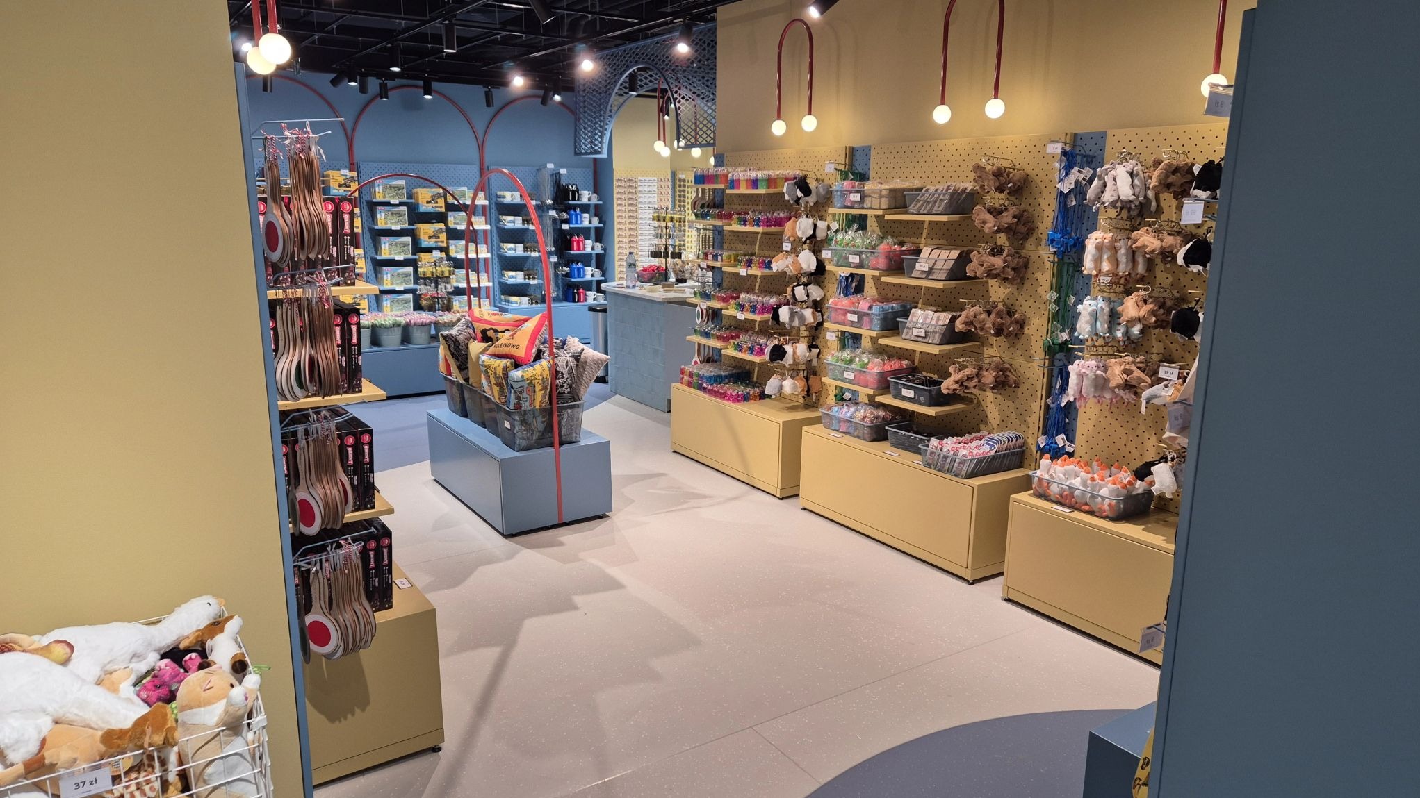 Wnętrze sklepu z artykułami dekoracyjnymi i upominkami, z ekspozycją towarów na półkach i w koszach. Widoczne oświetlenie i aranżacja przestrzeni.