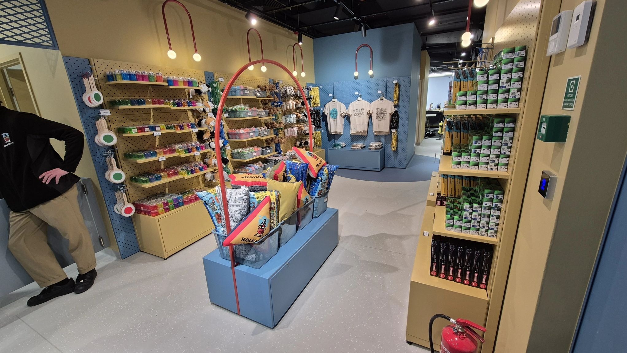 Wnętrze sklepu z pamiątkami: półki z zabawkami, poduszkami i koszulkami. Jasne oświetlenie, kolorowe wyposażenie, osoba w tle. Ujęcie z perspektywy klienta.