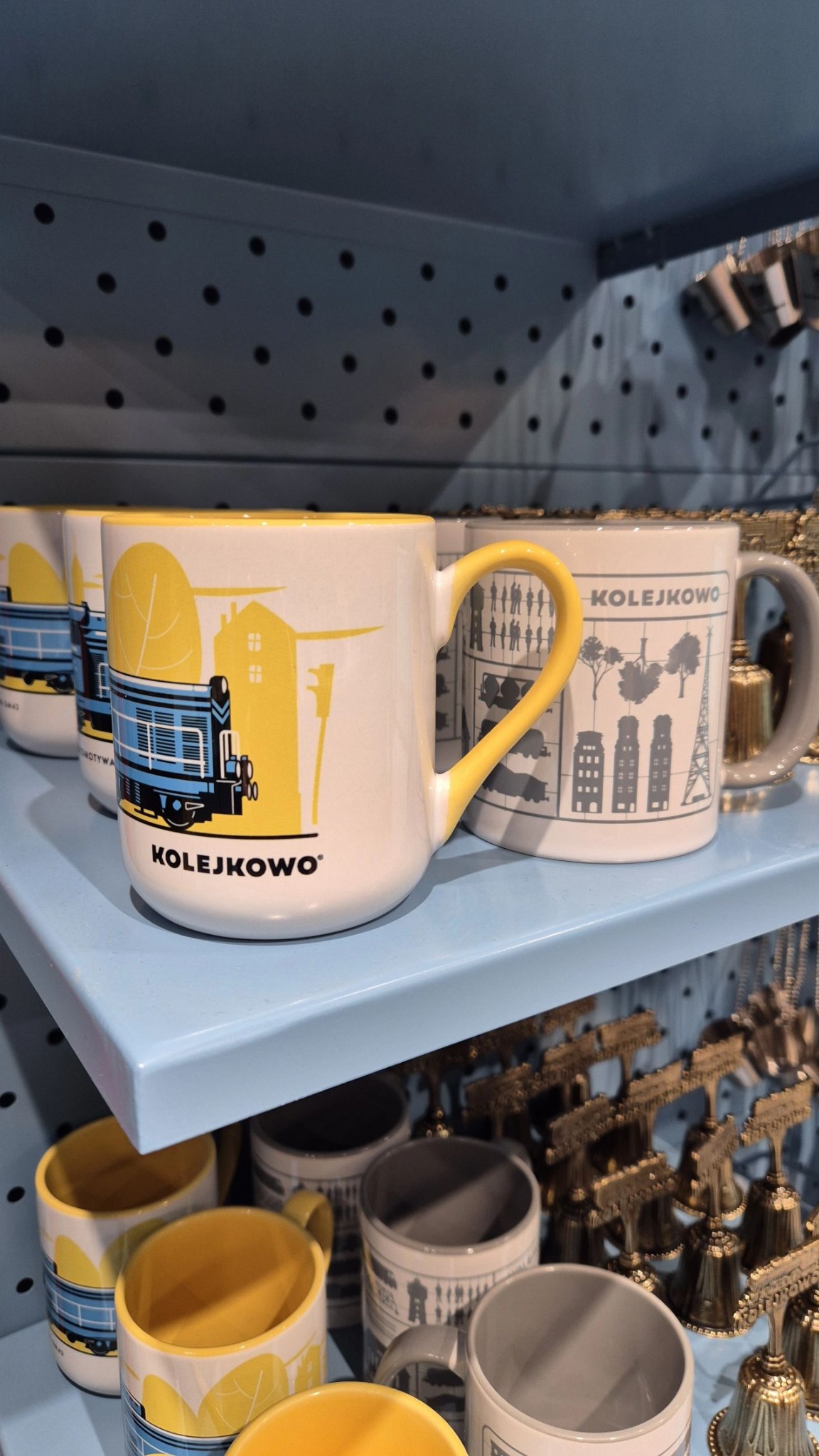 Kubki z motywem kolejowym i miejskim na błękitnej półce, z widocznym logo 'KOLEJKOWO'. Kubek z żółtym uchem i grafiką lokomotywy na pierwszym planie.