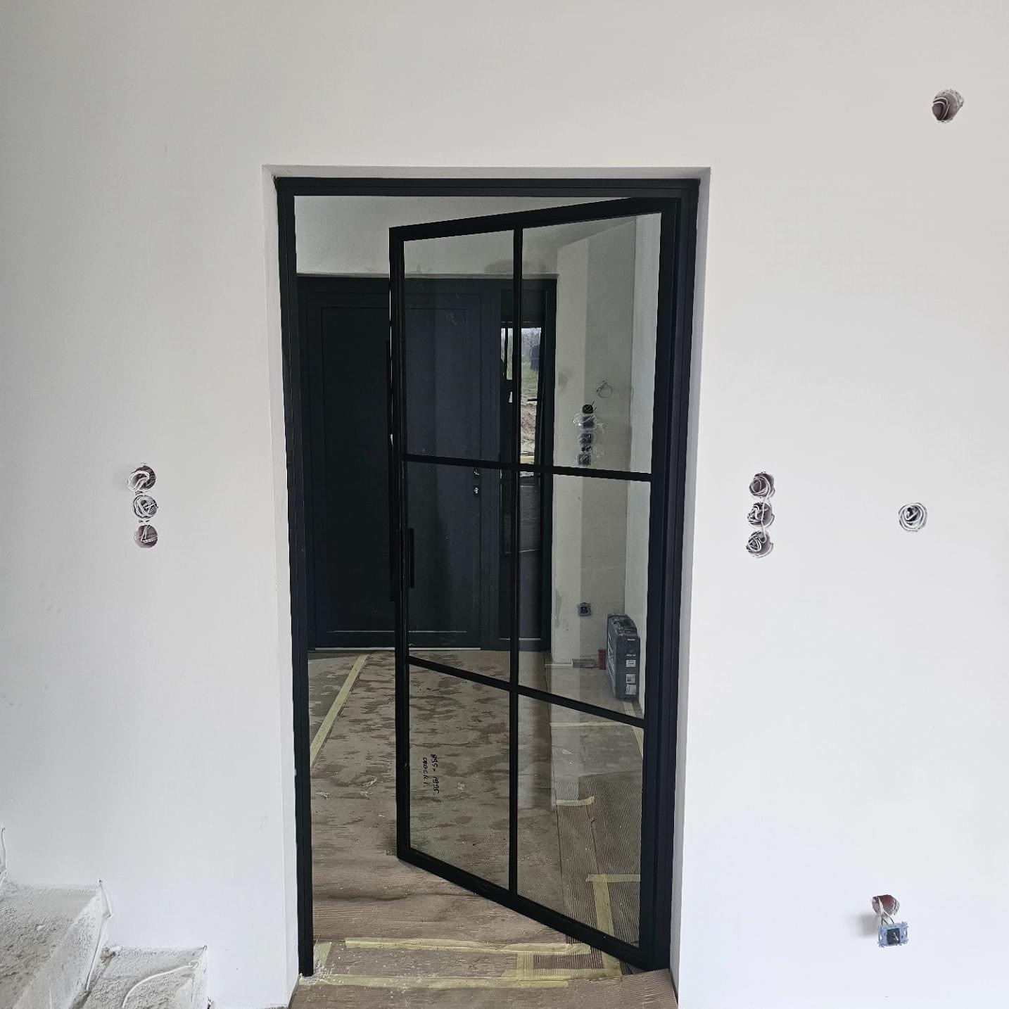 Loftowe drzwi wewnętrzne z czarną ramą i szprosami, częściowo otwarte, widoczne instalacje elektryczne na białej ścianie, surowy stan wykończenia wnętrza.