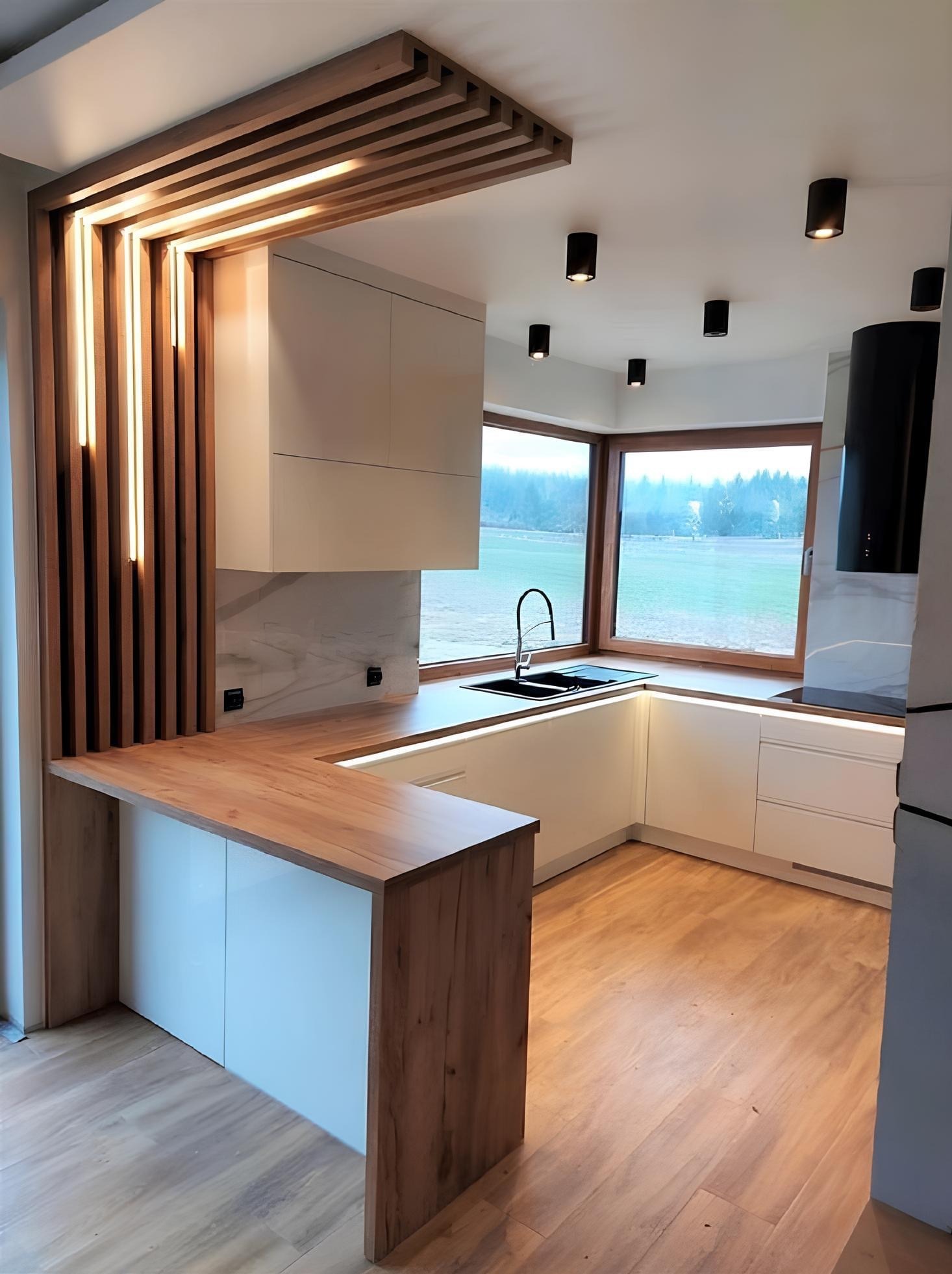 Nowoczesna kuchnia z drewnianą zabudową i oświetleniem LED, białe szafki, drewniany blat, okno z widokiem na zieleń. Minimalistyczny design, czarne lampy sufitowe.