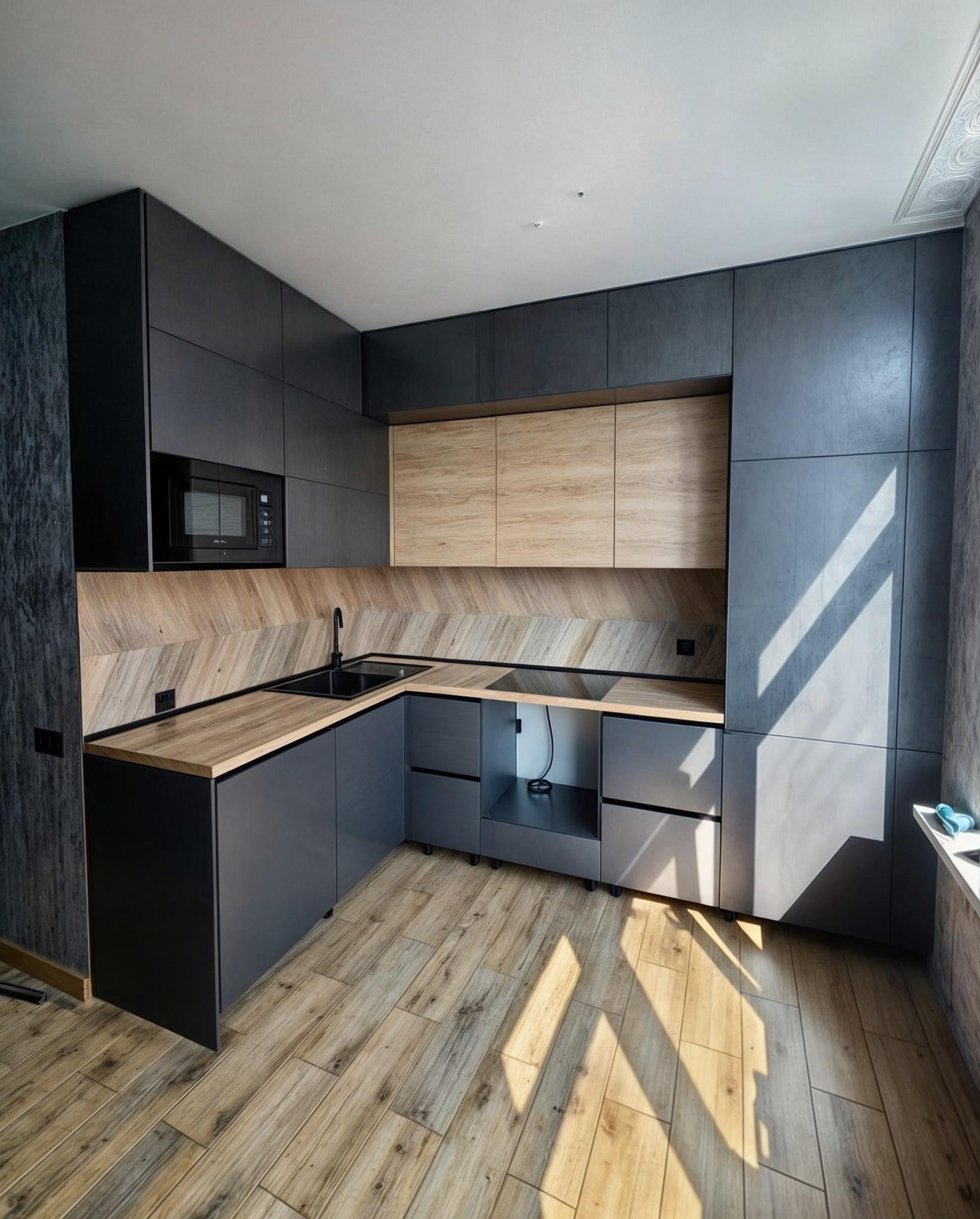 Nowoczesna kuchnia z ciemnymi, matowymi szafkami i drewnianymi akcentami, oświetlona promieniami słońca padającymi na drewnianą podłogę.