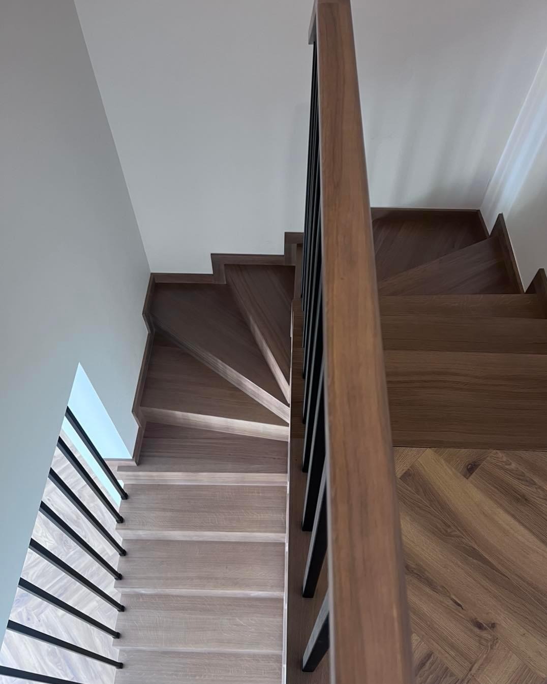 Drewniane schody z czarną, metalową balustradą, widok z góry. Stopnie wykończone parkietem w jodełkę, kontrastujące odcienie drewna. Minimalistyczny design.