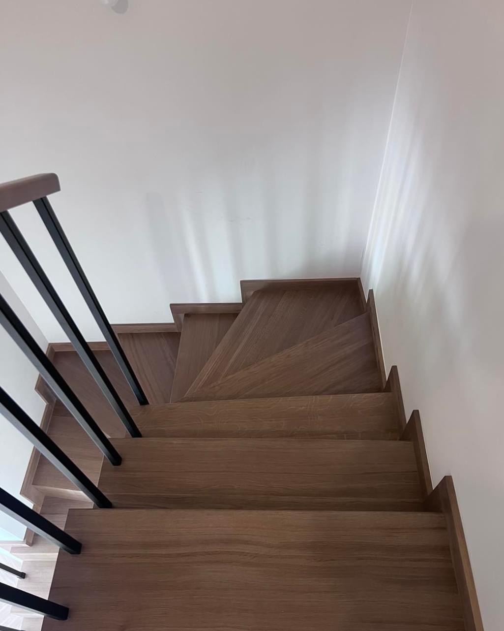 Kątowe ujęcie drewnianych schodów z czarną, minimalistyczną balustradą. Widok z góry podkreśla geometrię stopni i kontrast materiałów.