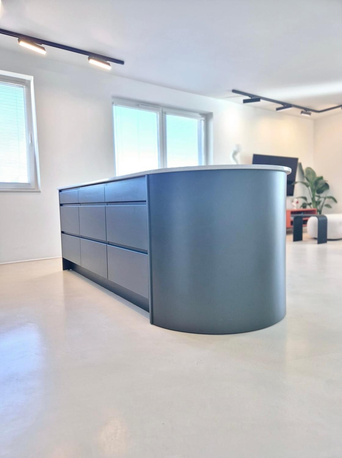 Nowoczesna wyspa kuchenna z szufladami w odcieniach szarości, zaokrąglony narożnik, blat z jasnego kamienia, minimalistyczne oświetlenie, jasna podłoga.