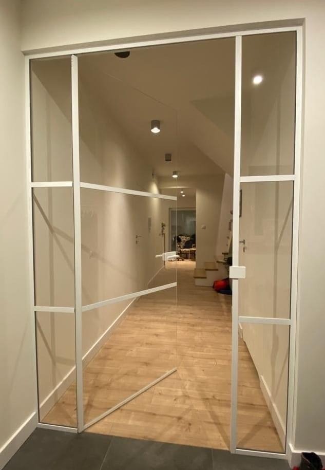 Biała, metalowa konstrukcja z szybami, tworząca przeszklone drzwi do korytarza z drewnianą podłogą. Widoczne oświetlenie sufitowe i schody w głębi.
