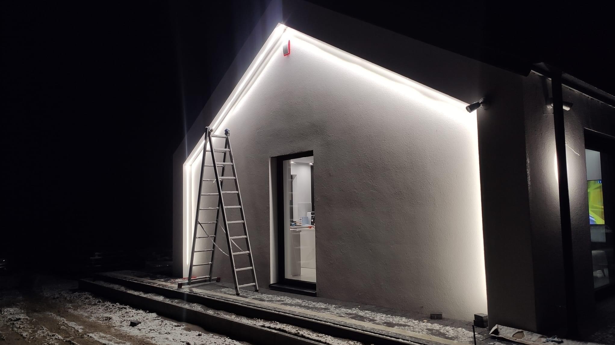 Oświetlenie elewacji budynku w nocy, drabina oparta o ścianę. Białe światło podkreśla architekturę. Widoczne lampy elewacyjne i fragment wnętrza przez okno.