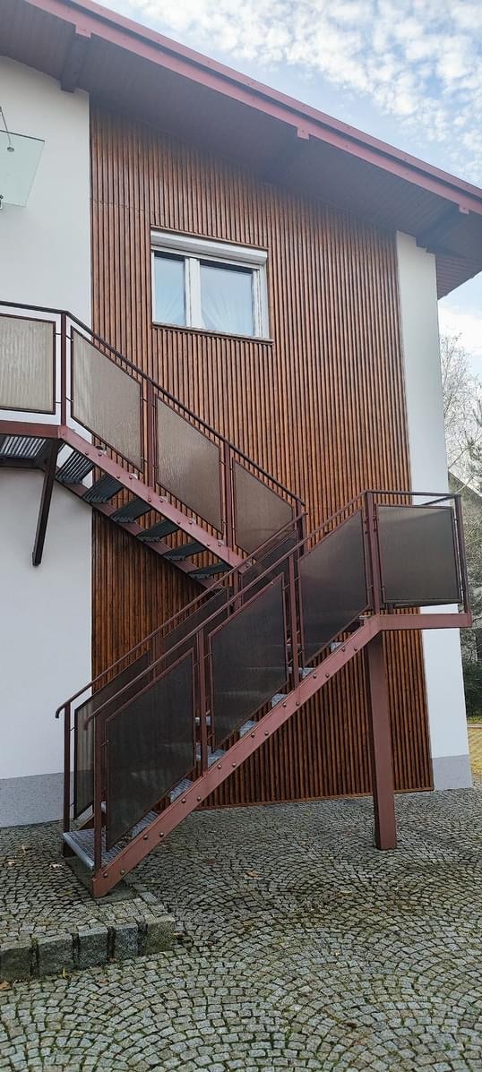 Zewnętrzne schody metalowe z brązowymi balustradami i wypełnieniem z przezroczystego tworzywa, zamontowane przy budynku z drewnianą elewacją.