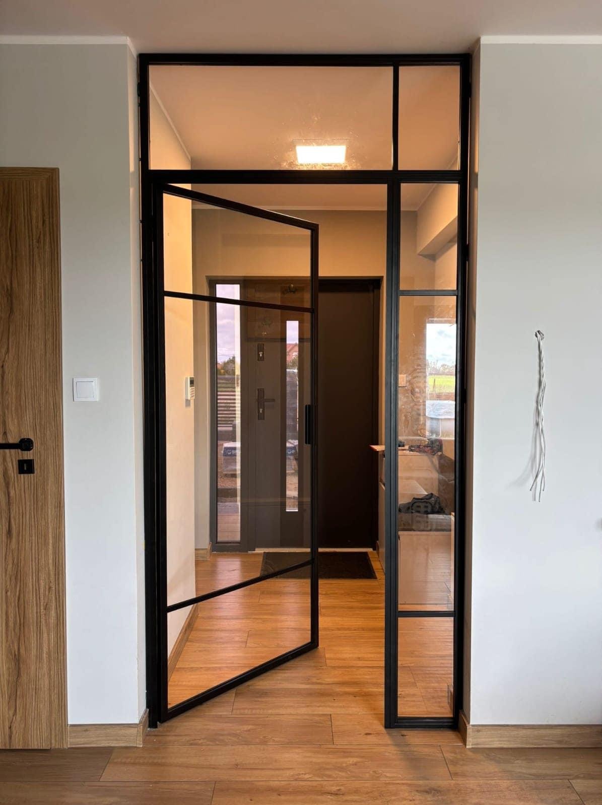 Nowoczesna, przeszklona ścianka działowa z czarną ramą, z otwartymi drzwiami, oddziela przestrzeń w domu. Widoczna podłoga z paneli i fragment ściany z kablem.