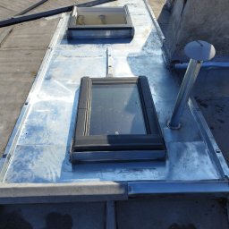 USŁUGI BUDOWLANE WOJCIECH WALA - Błyszczące, metalowe obróbki blacharskie dachu z dwoma oknami dachowymi i kominem wentylacyjnym. Widok z góry na dachy kamienic.