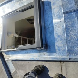 USŁUGI BUDOWLANE WOJCIECH WALA - Metalowa konstrukcja dachu z otworem, widok z góry na nogi pracownika w roboczych butach. Wewnątrz otworu widoczna drabina i elementy wyposażenia.