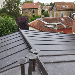 USŁUGI BUDOWLANE WOJCIECH WALA - Ciemny, metalowy dach z trzema kominami wentylacyjnymi na tle zabudowy miejskiej z dachami krytymi czerwoną dachówką. Widok z góry.