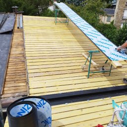 USŁUGI BUDOWLANE WOJCIECH WALA - Dach z drewnianą konstrukcją, częściowo pokryty papą. Widoczne narzędzia dekarskie i rolka papy. W tle drzewa i budynki.