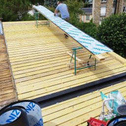 USŁUGI BUDOWLANE WOJCIECH WALA - Montaż blachy na dachu z drewnianą konstrukcją. Widoczne narzędzia i materiały budowlane, dekarz w trakcie pracy. Tło stanowią budynki i zieleń.