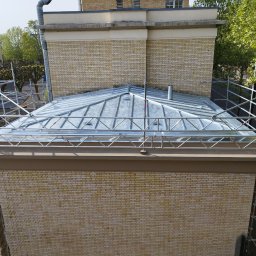 USŁUGI BUDOWLANE WOJCIECH WALA - Jasna, blaszana konstrukcja dachu budynku z cegły, widziana z góry. Na dachu widoczne rusztowania. W tle budynek z kominem i drzewa.