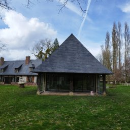 USŁUGI BUDOWLANE WOJCIECH WALA - Widok na dom z dachem z ciemnej dachówki i nową konstrukcją z przeszklonymi ścianami, również z dachem z dachówki, na tle zielonego trawnika i błękitnego nieba.