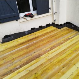 USŁUGI BUDOWLANE WOJCIECH WALA - Fragment dachu z żółtych desek, zabezpieczony czarną folią izolacyjną. Na parapecie leżą narzędzia dekarskie: zszywacz i młotek. Widoczne okno z niebieskimi okiennicami.