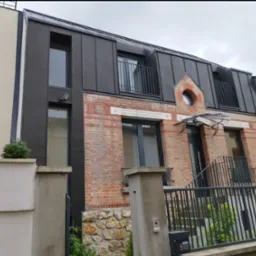 Dom z czarnym dachem z blachy, ceglastą elewacją i balkonami. Uwagę zwraca okrągłe okno na poddaszu i nowoczesne wykończenie.