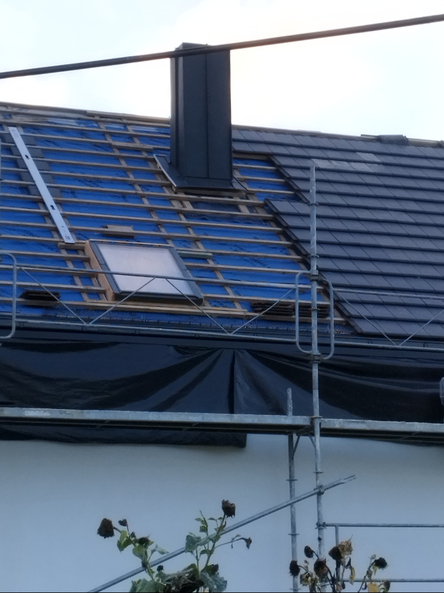 Dach w trakcie krycia, widoczne rusztowanie, okno dachowe i komin. Część dachu pokryta dachówką, a część z niebieską membraną i łatami. Ujęcie z dołu.