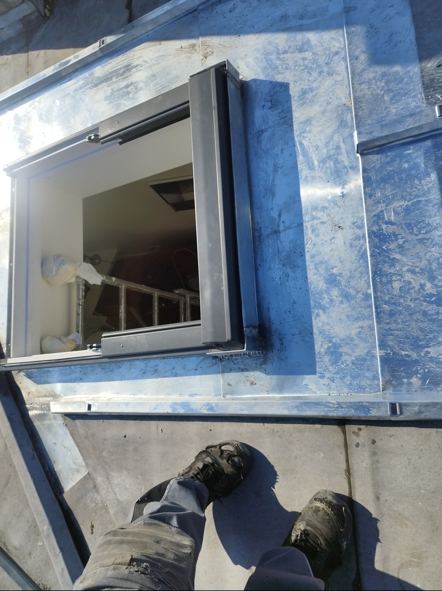 Metalowa konstrukcja dachu z otworem, widok z góry na nogi pracownika w roboczych butach. Wewnątrz otworu widoczna drabina i elementy wyposażenia.