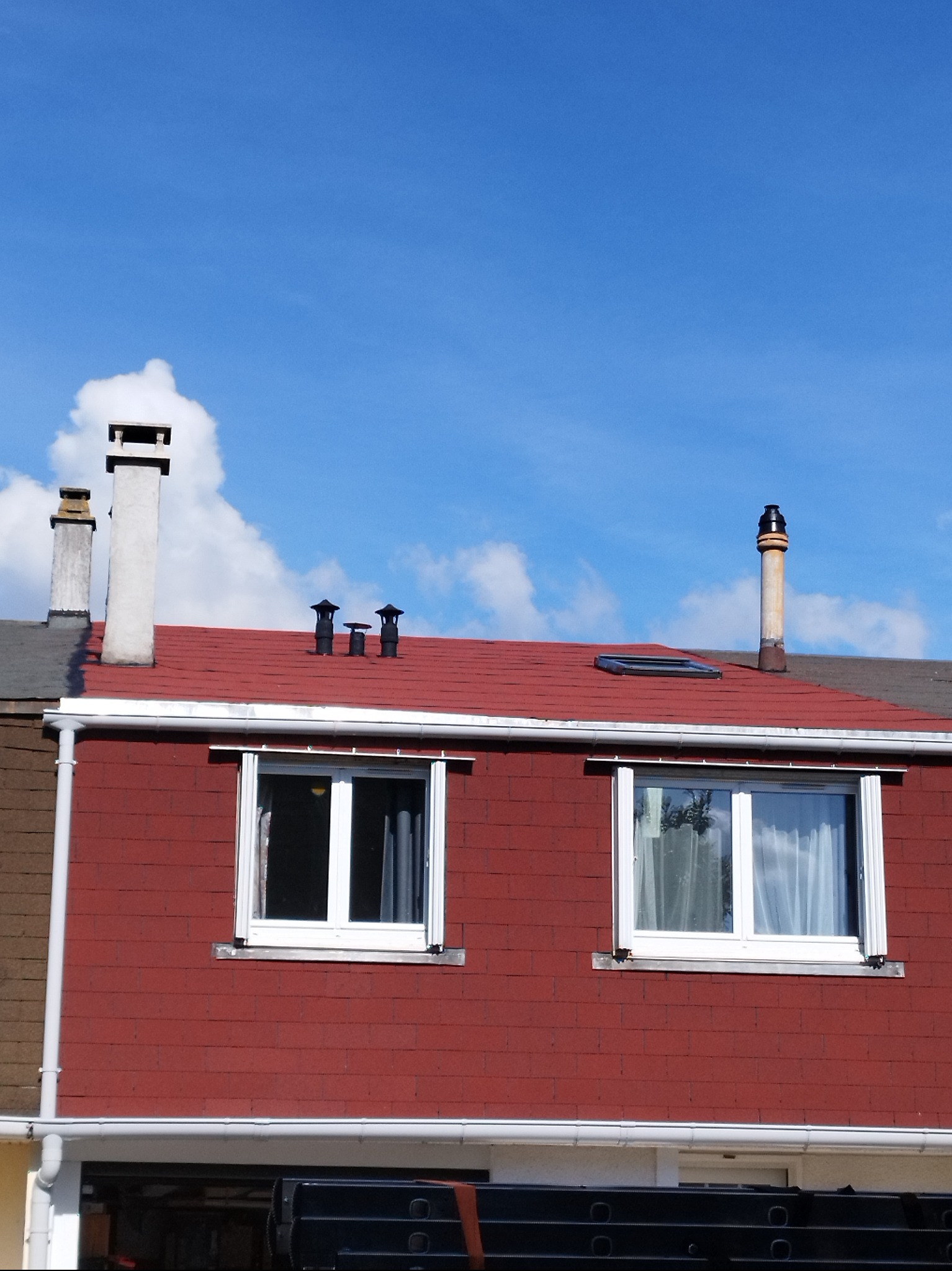 Czerwony dach z kilkoma kominami na tle błękitnego nieba z chmurami. Dwa okna z białymi ramami. Widoczny fragment rynny i drabiny.