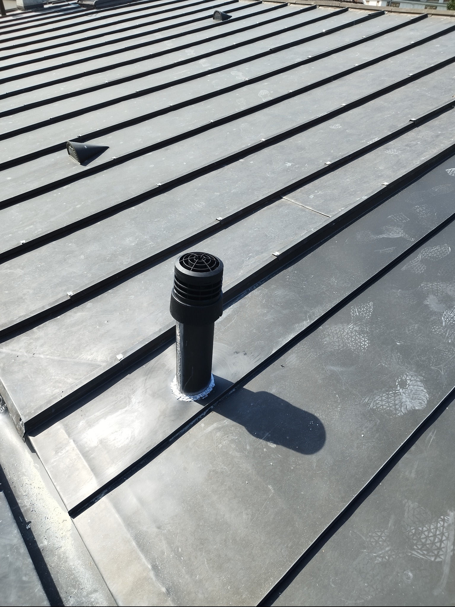 Szary dach z blachy na rąbek stojący z kominkiem wentylacyjnym. Widoczne ślady butów. Ujęcie z góry, w słoneczny dzień.