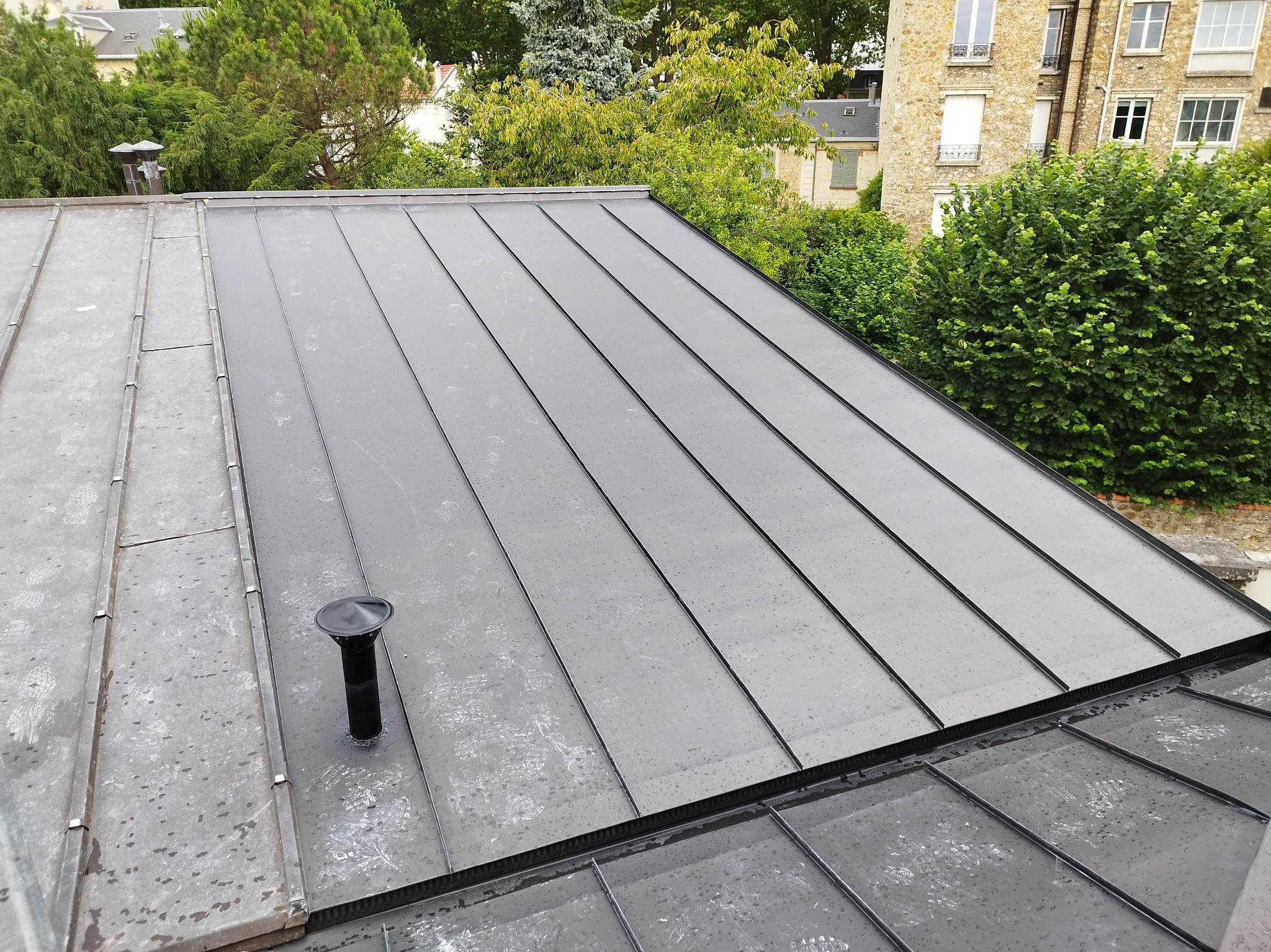 Nowoczesny, grafitowy dach z blachy na tle zieleni i kamienicy. Widoczny kominek wentylacyjny. Minimalistyczne wykończenie, regularne linie. Dach po deszczu.