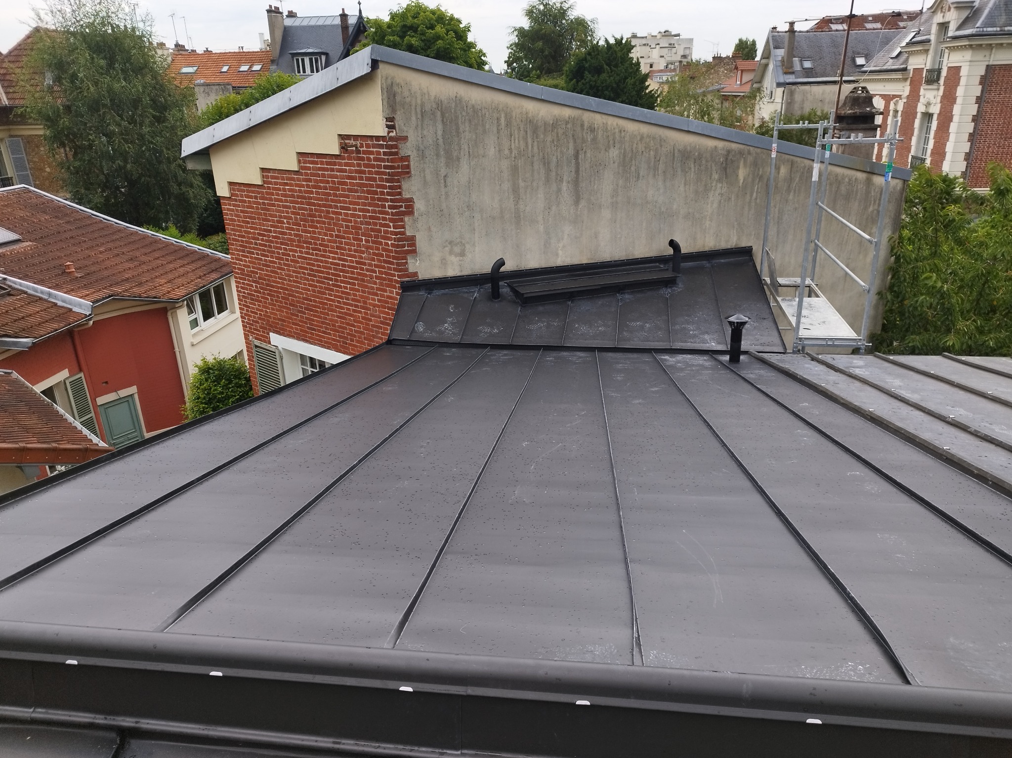 Nowy, czarny dach z blachy na budynku miejskim, widok z góry. Widoczne kominy i elementy obróbek blacharskich. W tle zabudowa miejska i zieleń.