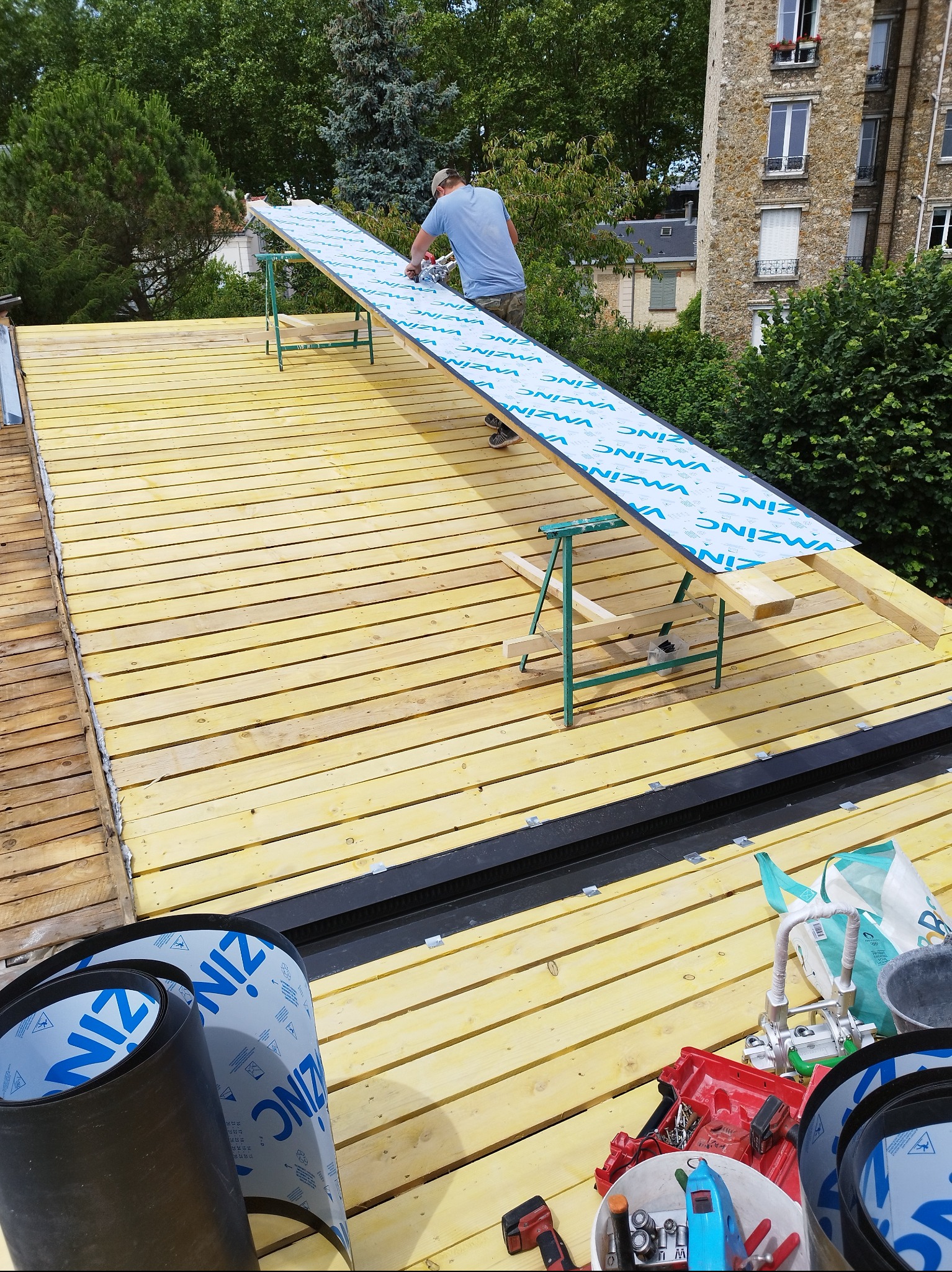 Montaż blachy na dachu z drewnianą konstrukcją. Widoczne narzędzia i materiały budowlane, dekarz w trakcie pracy. Tło stanowią budynki i zieleń.