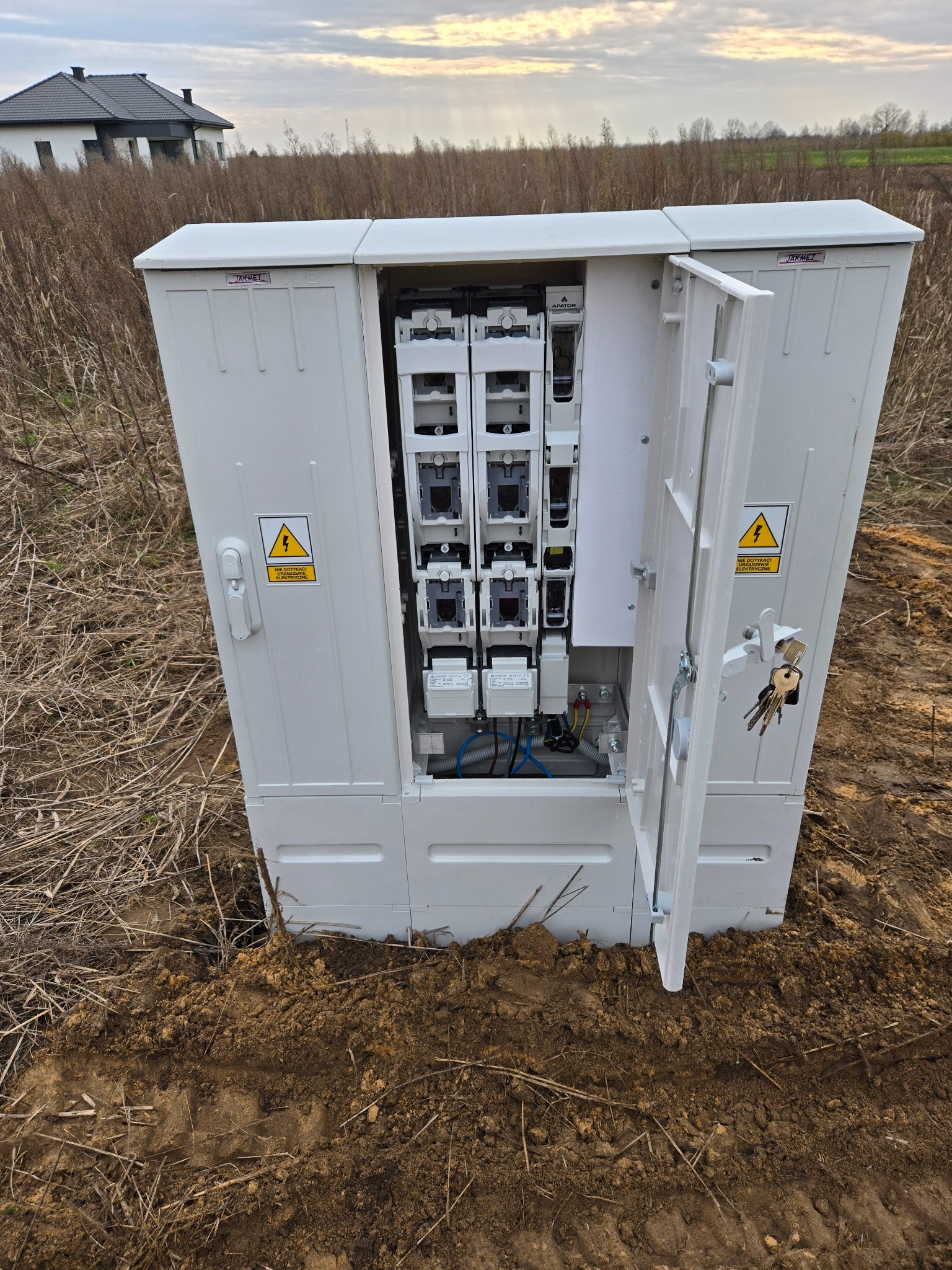 Otwarta, biała skrzynka elektryczna z bezpiecznikami na zewnątrz, na tle pola i domu. Widoczne kable i elementy instalacji wewnątrz. Klucze wiszą na drzwiczkach.