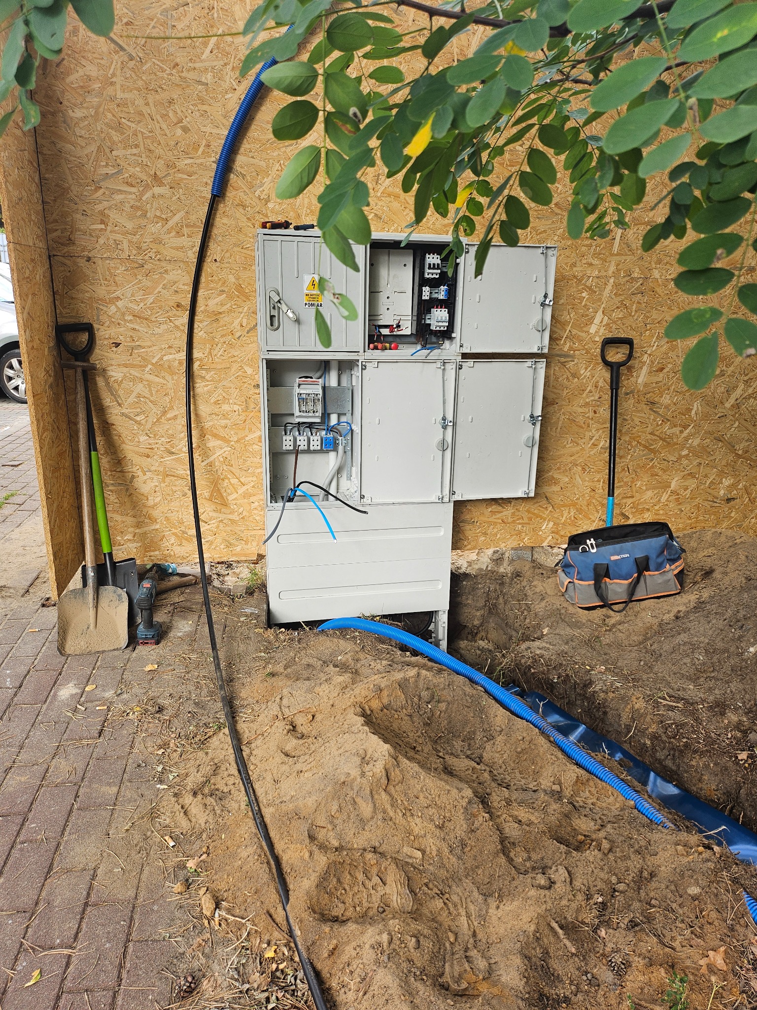 Otwarta skrzynka elektryczna z podłączonymi przewodami, narzędzia pracy (szpadle, wiertarka, torba narzędziowa) obok wykopu. Widok z bliska, oświetlenie dzienne.