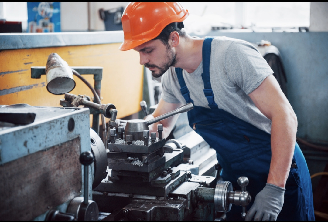 Pracownik w kasku obsługuje tokarkę w warsztacie. Metalowe wióry na maszynie, lampa oświetla obszar roboczy. Mężczyzna w kombinezonie i rękawicach ochronnych.