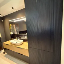 Nowoczesna łazienka z umywalką nablatową, drewnianą szafką i wysoką, czarną szafą z pionowymi żłobieniami. Lustro z oświetleniem LED.