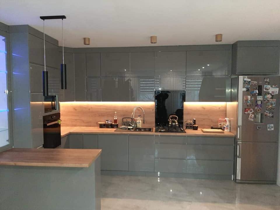 Szara kuchnia z połyskiem, drewniany blat, podświetlenie LED pod szafkami, nowoczesne lampy wiszące. Lodówka z magnesami po prawej stronie.