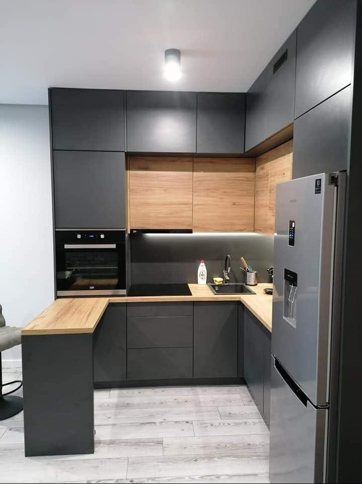 Nowoczesna kuchnia z szarymi szafkami i drewnianymi blatami, oświetleniem LED pod szafkami wiszącymi i srebrną lodówką. Minimalistyczny design.