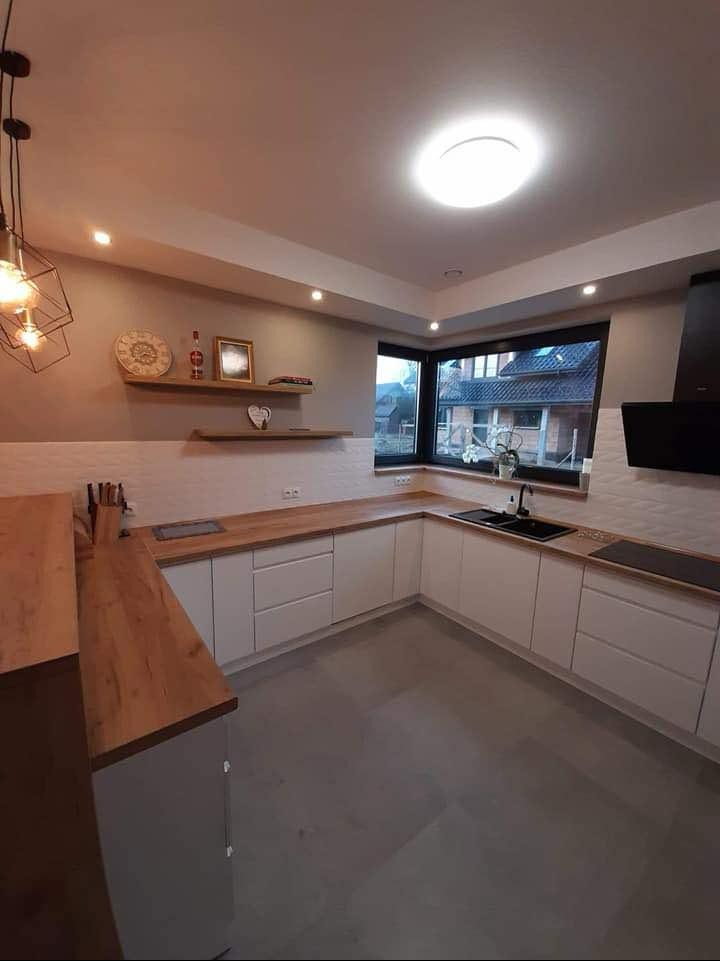 Jasna kuchnia w kształcie litery U z drewnianymi blatami i białymi frontami, okno z widokiem na dom, nowoczesne oświetlenie.