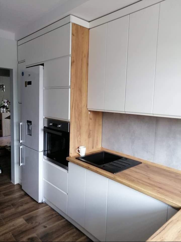 Biała kuchnia z drewnianym blatem i zabudową, z czarnym zlewem i białą filiżanką. Widoczna lodówka i piekarnik. Nowoczesny design, drewnopodobne panele na podłodze.