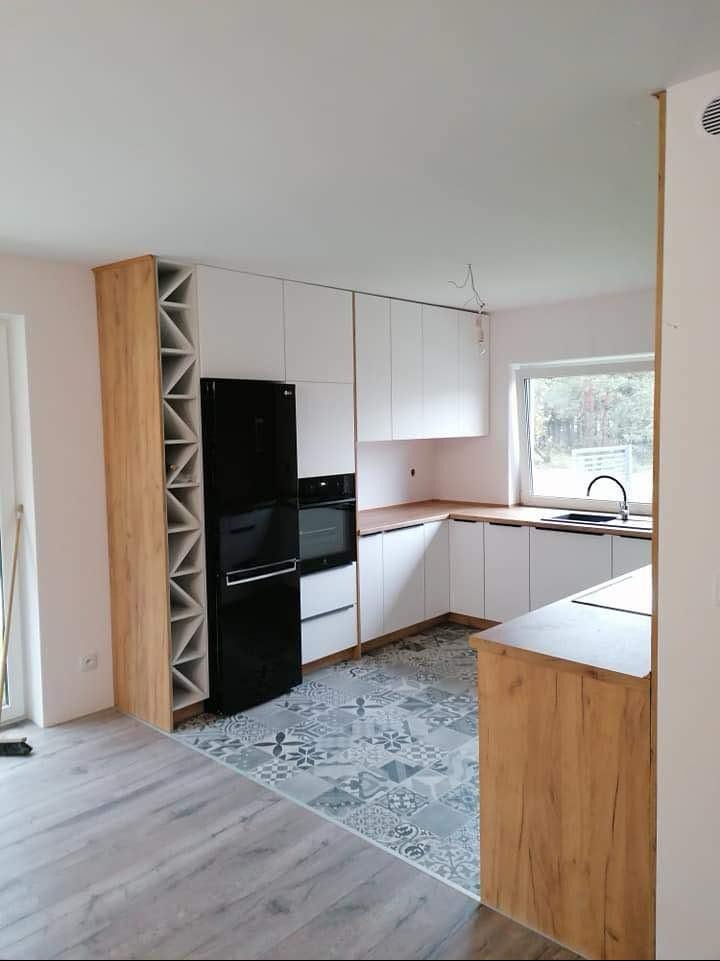 Nowoczesna kuchnia z białymi szafkami i drewnianymi akcentami. Uwagę przykuwa regał na wino w kolorze drewna obok czarnej lodówki. Na podłodze wzorzyste płytki.