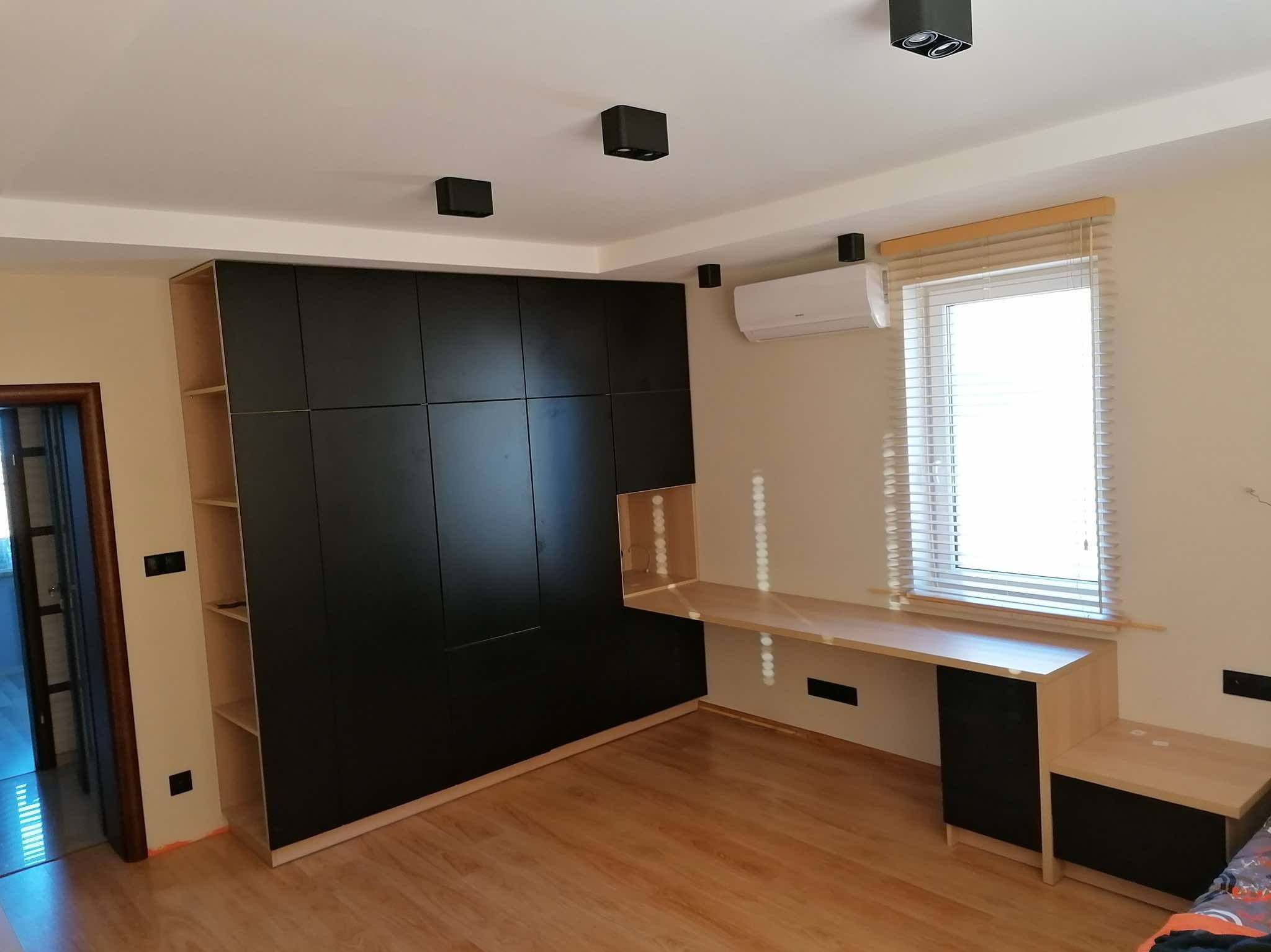 Czarna szafa wnękowa z biurkiem w jasnym pokoju. Widoczne drewniane półki, roleta w oknie i lampy sufitowe. Nowoczesny design, minimalistyczne wykończenie.