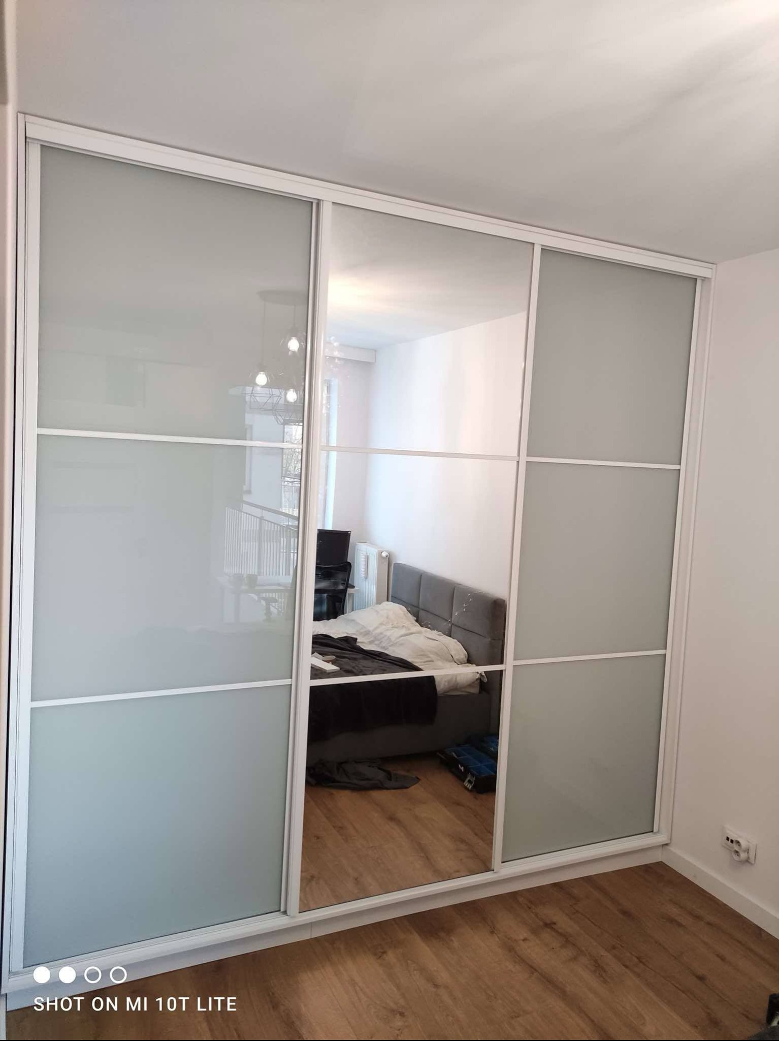 Biała szafa wnękowa z przesuwnymi drzwiami: jedna z lustrem odbija wnętrze pokoju z łóżkiem, pozostałe z mlecznego szkła, z podziałem na sześć kwadratów.