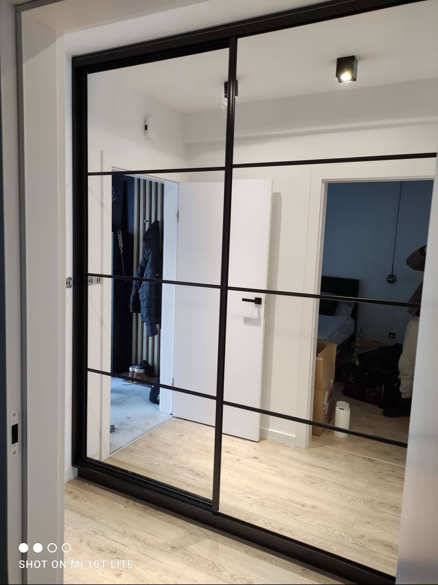 Nowoczesna szafa wnękowa z lustrzanymi drzwiami przesuwnymi i czarnymi szprosami, odbijająca wnętrze mieszkania z drewnianą podłogą i elementami dekoracyjnymi.