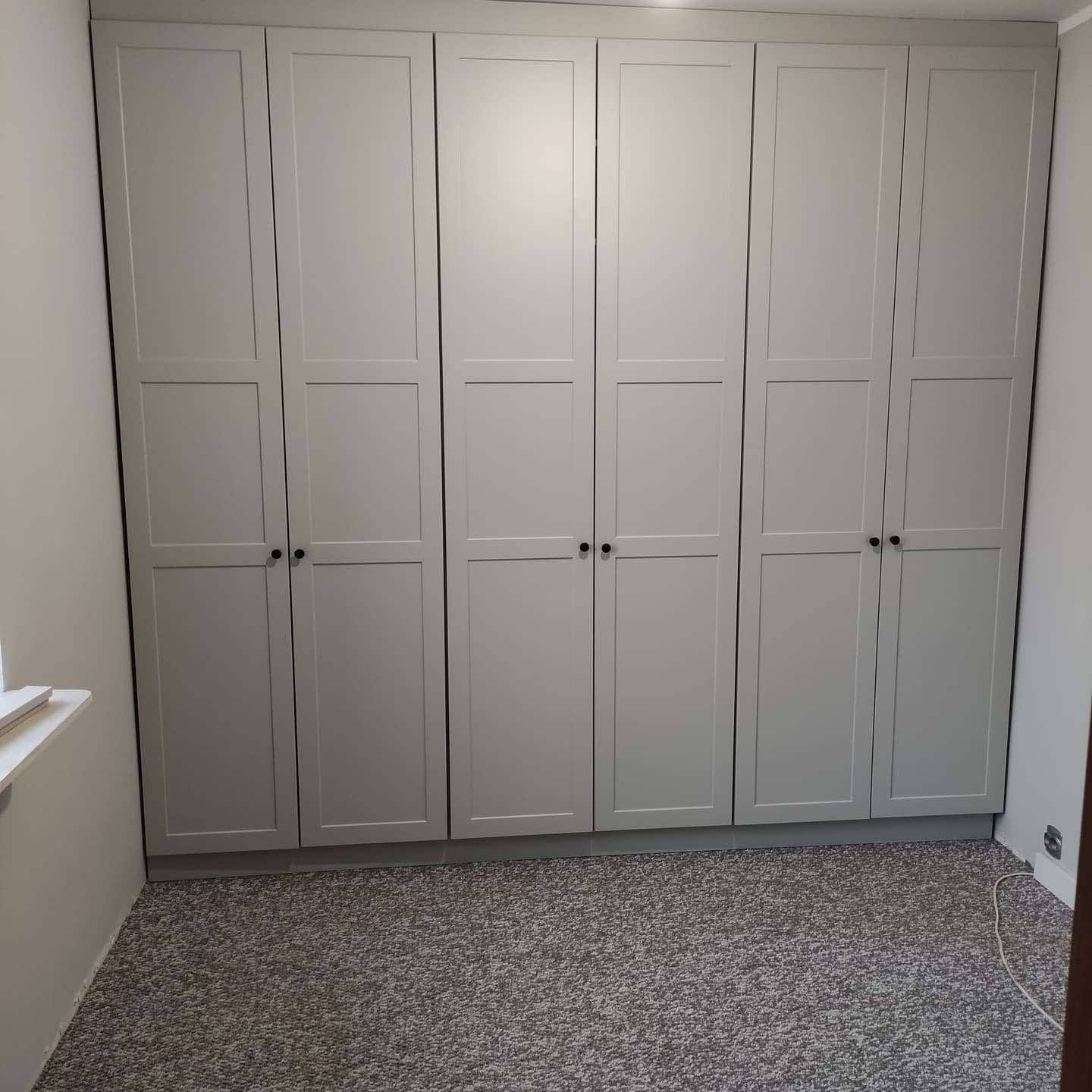 Jasnoszara szafa wnękowa z sześcioma drzwiami z panelami i czarnymi uchwytami, dopasowana do podłogi wyłożonej wykładziną dywanową w pokoju.