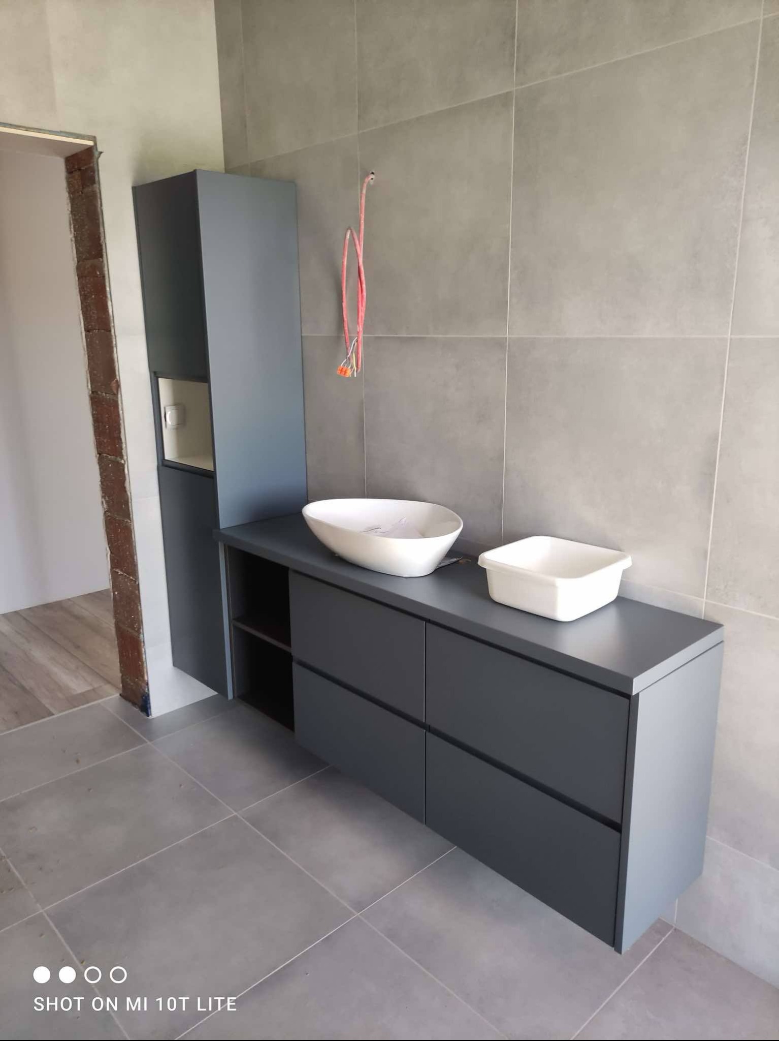 Nowoczesna szara szafka łazienkowa z dwiema umywalkami na blacie, obok wysoka szafka, szare płytki na ścianie i podłodze, widoczne kable ze ściany.