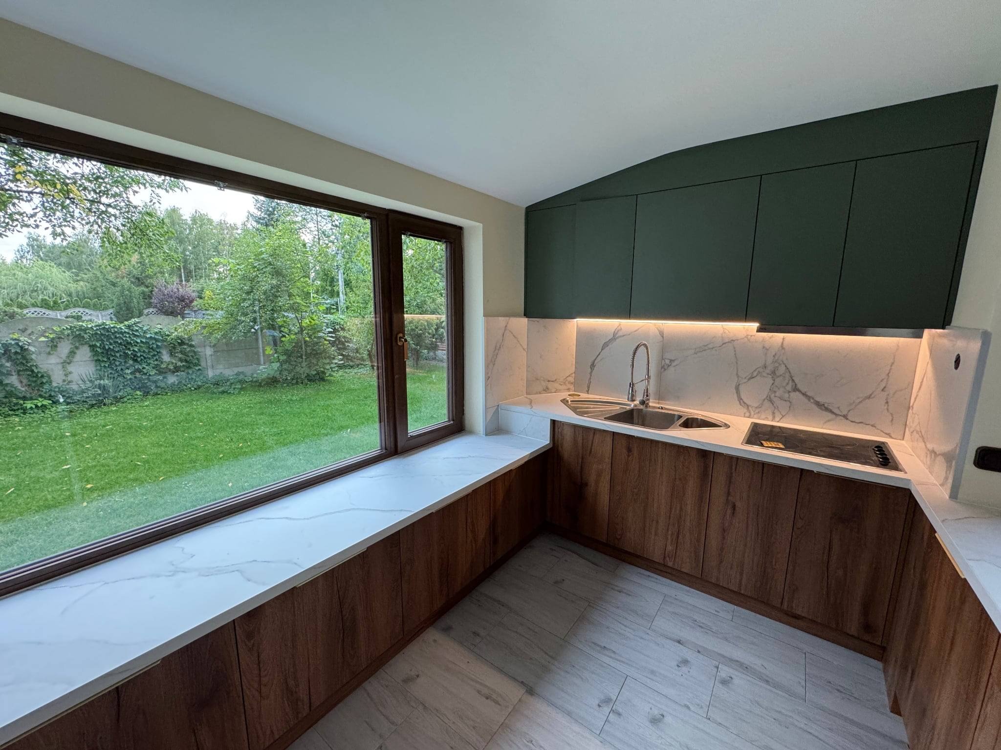 Nowoczesna kuchnia z drewnianymi szafkami, białym blatem z marmurowym wzorem i dużym oknem z widokiem na zieleń. Górne szafki w kolorze ciemnej zieleni.